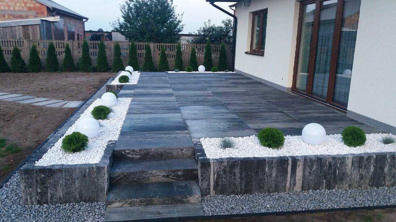 Wykończony taras z kostki brukowej w odcieniach szarości, z niskimi schodami, obrzeżami z białego kamienia i kulistymi lampami ogrodowymi, na tle domu z brązowymi oknami i zielonych tuj.