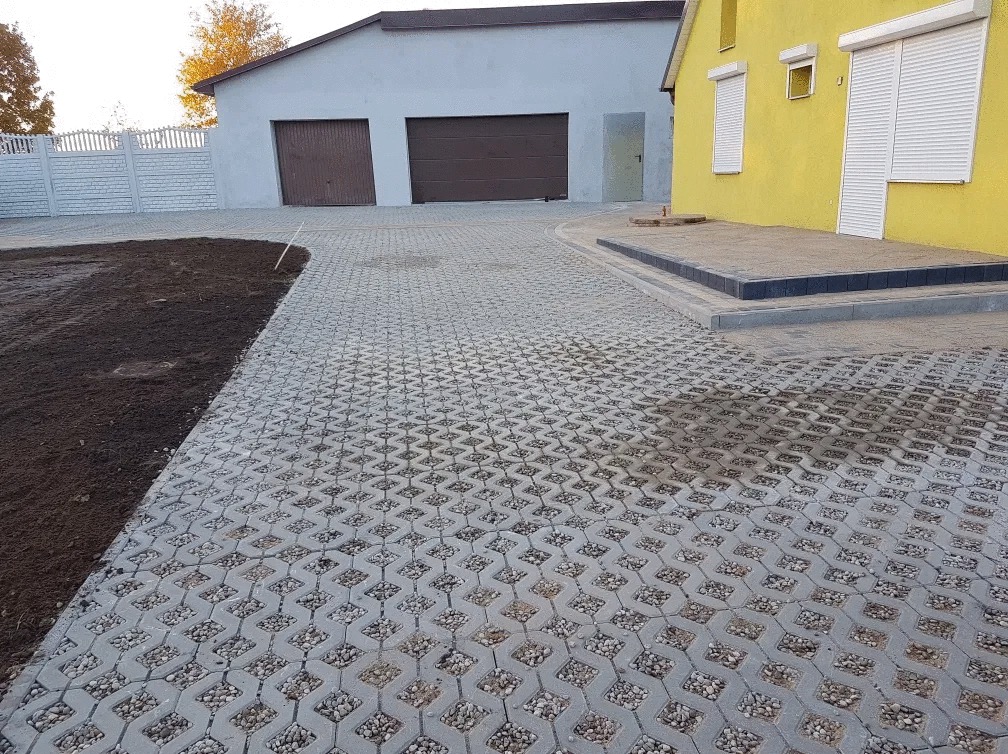 Szara ażurowa nawierzchnia z wypełnieniem z kamieni, prowadząca do żółtego domu z białymi okiennicami i szarego garażu, świeżo przygotowany teren pod trawnik po lewej stronie.