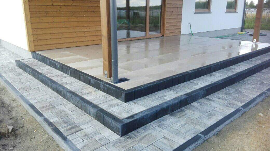 Schody wejściowe do domu wyłożone szarą kostką brukową z ciemnymi obrzeżami, prowadzące do tarasu z jasnych płyt, widoczny fragment drewnianej elewacji i okna.