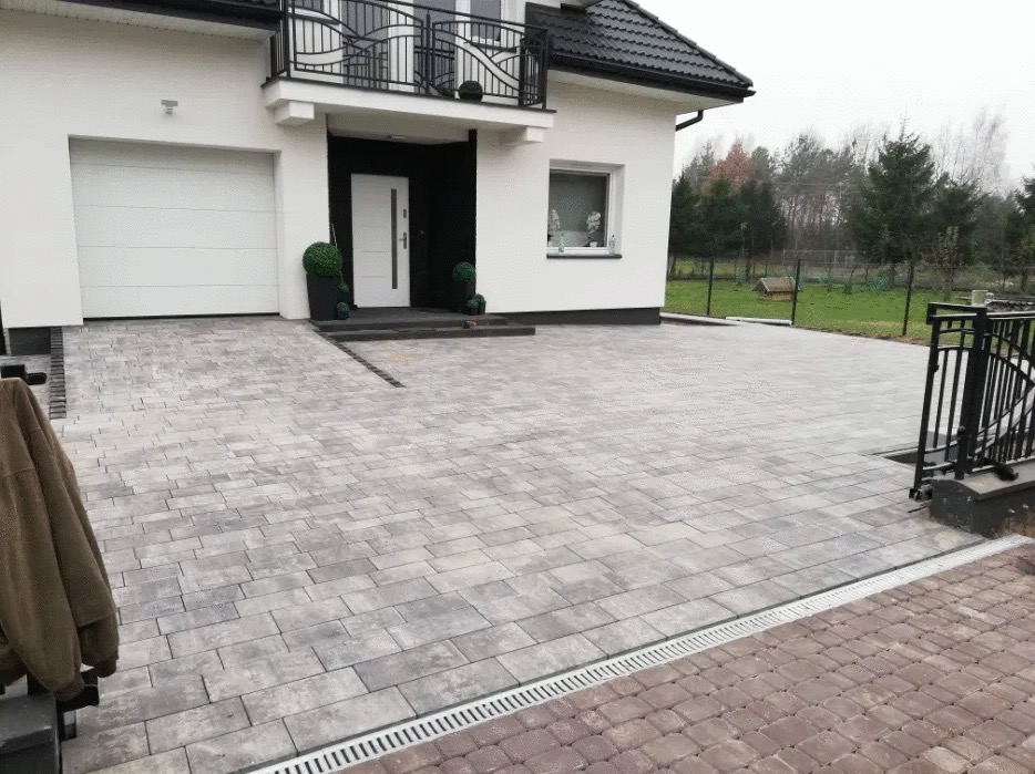 Ułożona szara kostka brukowa na podjeździe przed nowoczesnym domem z białymi ścianami i czarnym dachem, widoczny system odprowadzania wody.