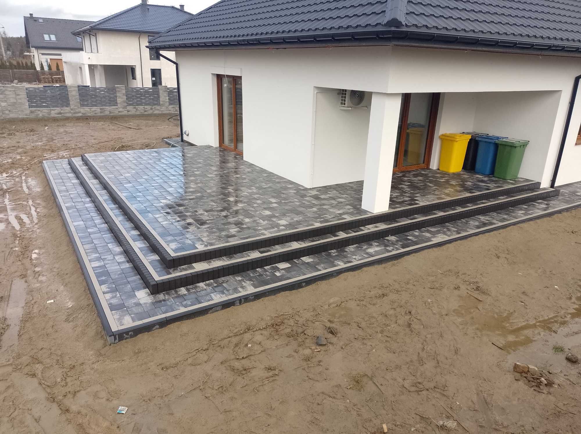 Nowoczesny taras z kostki brukowej w odcieniach szarości, z trzema stopniami i obrzeżami, przylegający do białego domu z widocznymi pojemnikami na śmieci i błotnistym terenem wokół.