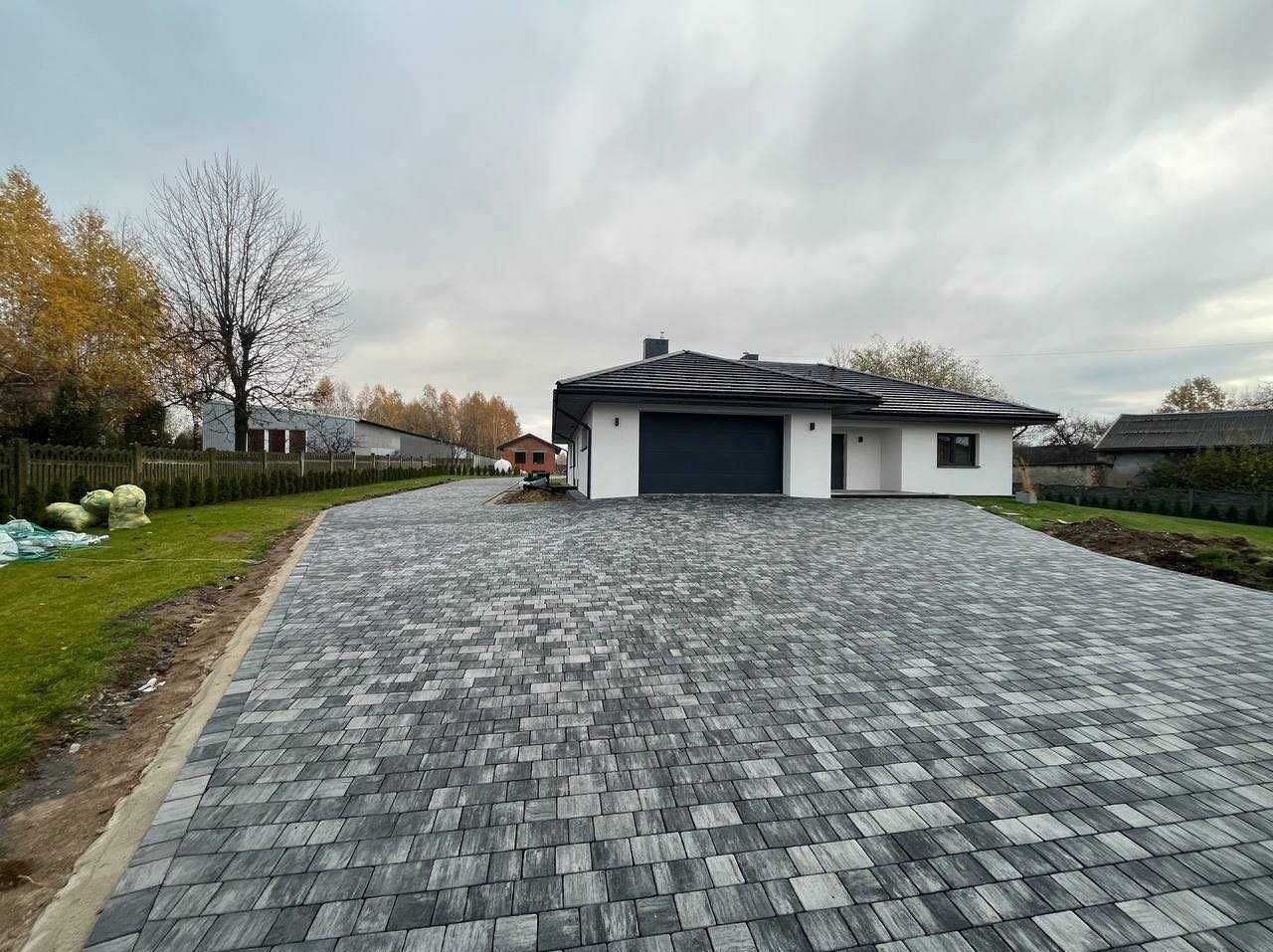 Nowoczesny dom z podjazdem wyłożonym szarą kostką w jodełkę; widok z perspektywy szerokokątnej, pochmurne niebo.