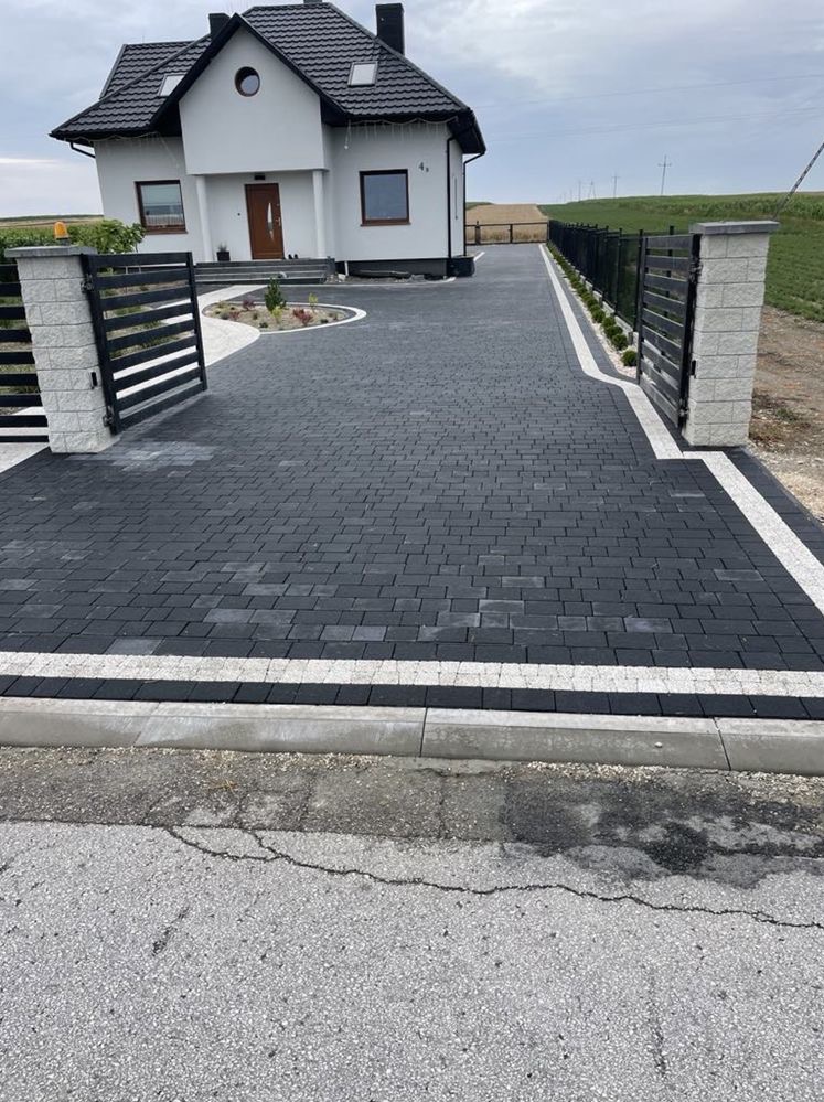 Czarna kostka brukowa ułożona na podjeździe do domu jednorodzinnego, obrzeża z jasnej kostki, brama wjazdowa z elementami murowanymi.