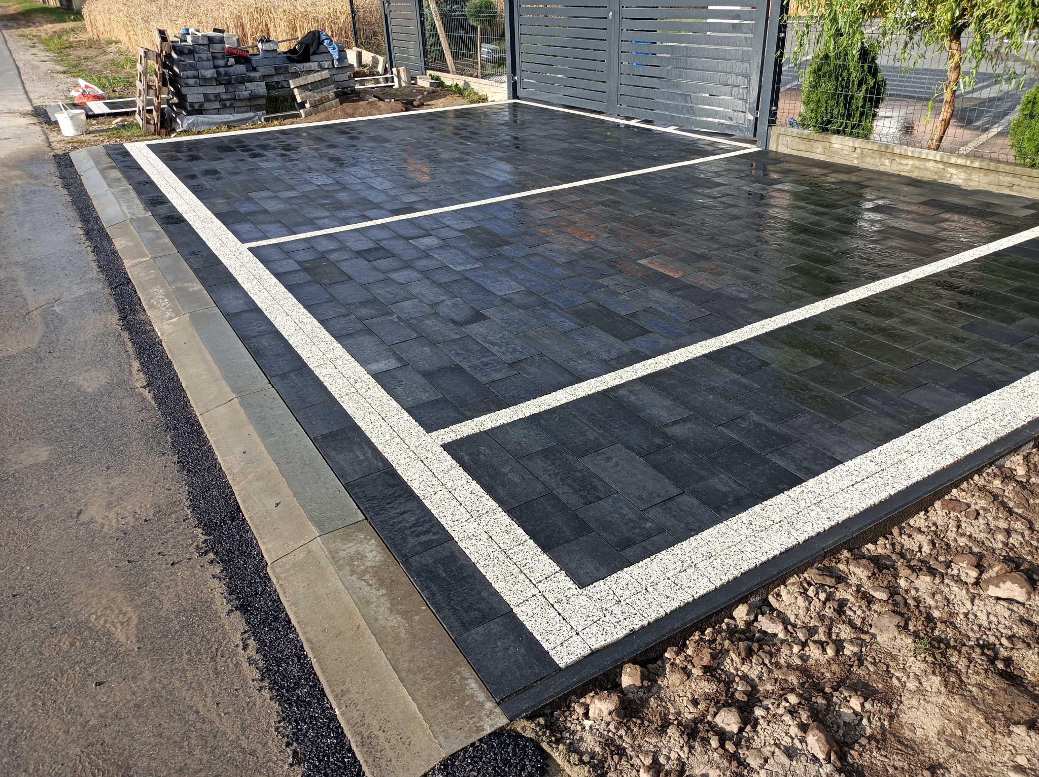 Ułożona, czarna kostka brukowa z białymi, dekoracyjnymi pasami, tworząca wzór prostokątów na podjeździe, widoczne obrzeża i resztki ziemi po pracach.