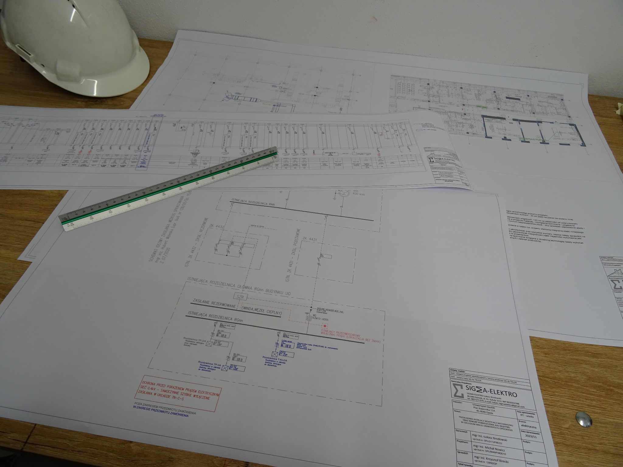 Rozłożone na drewnianym stole plany instalacji elektrycznych, linijka z podziałką centymetrową i biały kask ochronny. Widoczny schemat ideowy zasilania i logo firmy SIGMA-ELEKTRO.
