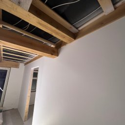 Dam-bud - Wnętrze w trakcie prac wykończeniowych: świeżo pomalowana ściana w jasnym odcieniu szarości, widoczne drewniane elementy konstrukcyjne sufitu oraz przygotowane otwory drzwiowe i okienne.