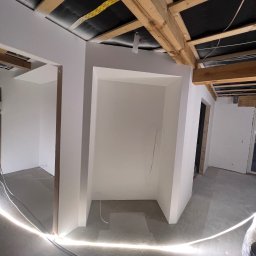 Dam-bud - Wnętrze w trakcie wykańczania: świeżo pomalowane na biało ściany z drewnianą konstrukcją sufitu, widoczne kable i przewody, oświetlenie podłogowe.