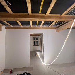 Dam-bud - Wnętrze w trakcie remontu: świeżo pomalowane białe ściany, odsłonięty drewniany strop z widoczną instalacją elektryczną, przewód LED na ścianie, sprzęt malarski w rogu, puszki po napojach na podłodze.