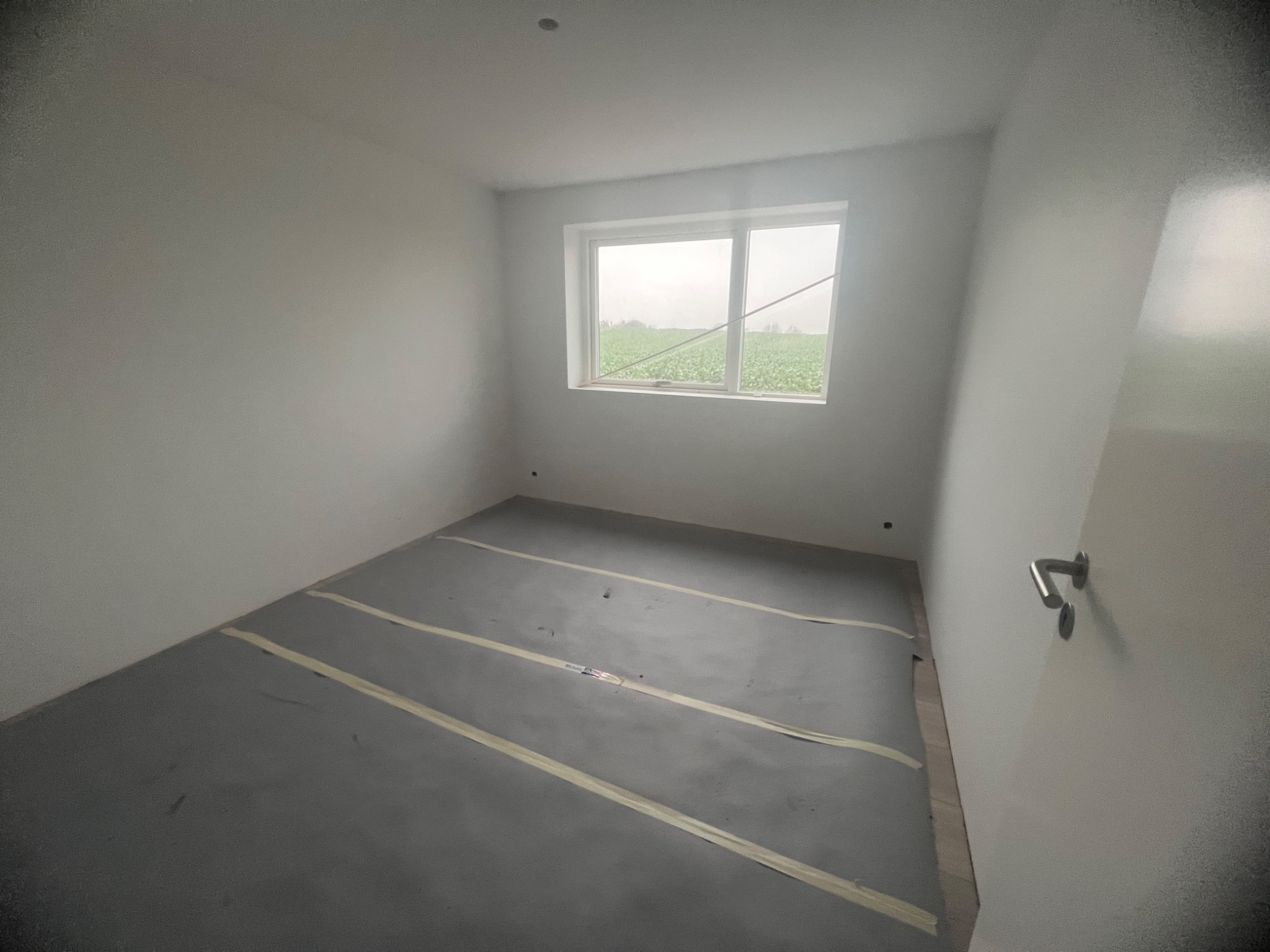 Przygotowane do malowania wnętrze pokoju z podłogą zabezpieczoną folią i taśmą malarską, widok na okno z zielenią na zewnątrz.