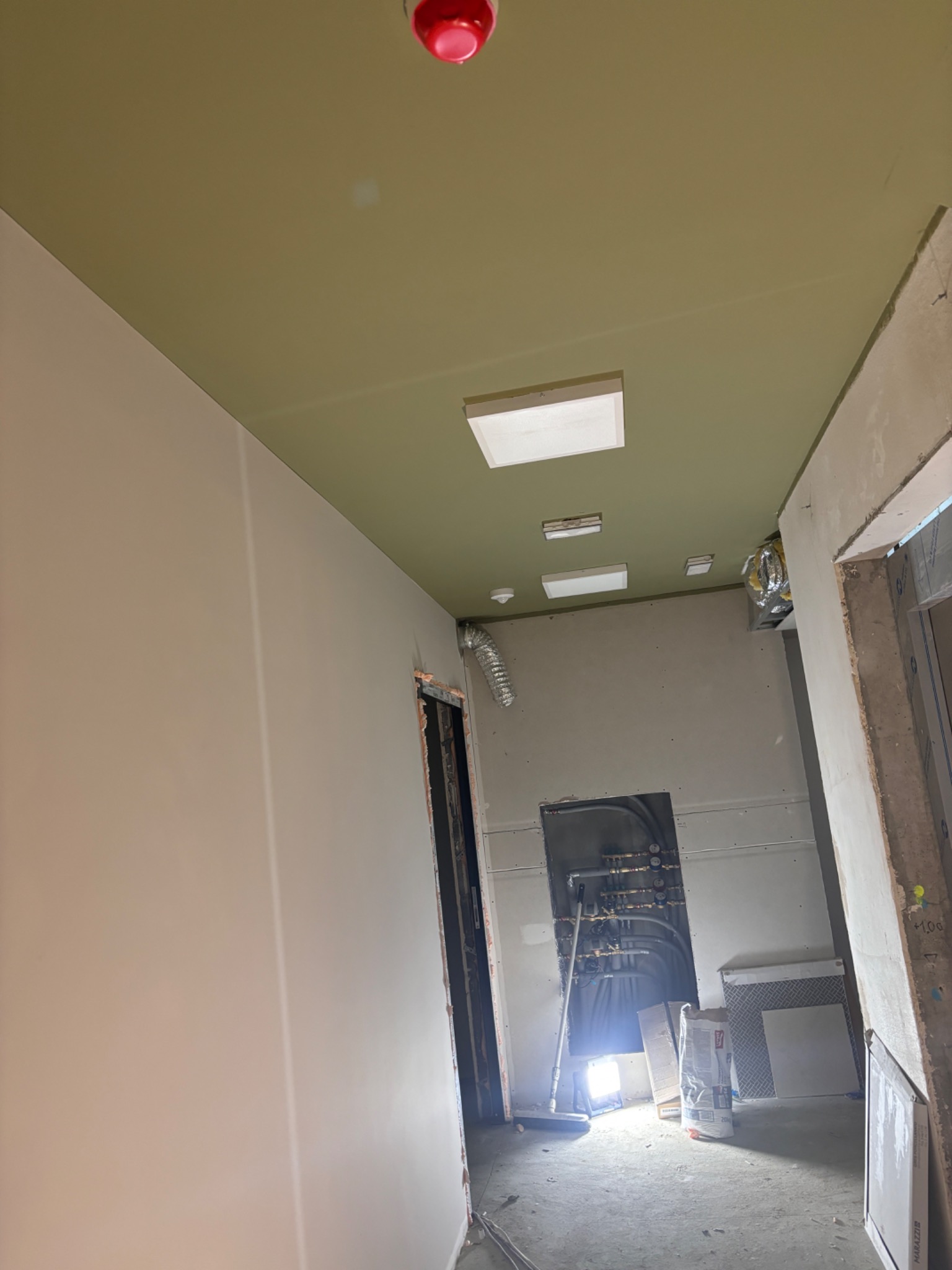 Korytarz w trakcie prac wykończeniowych: ściany przygotowane pod malowanie, sufit pomalowany na oliwkowy kolor, widoczne instalacje i materiały budowlane.