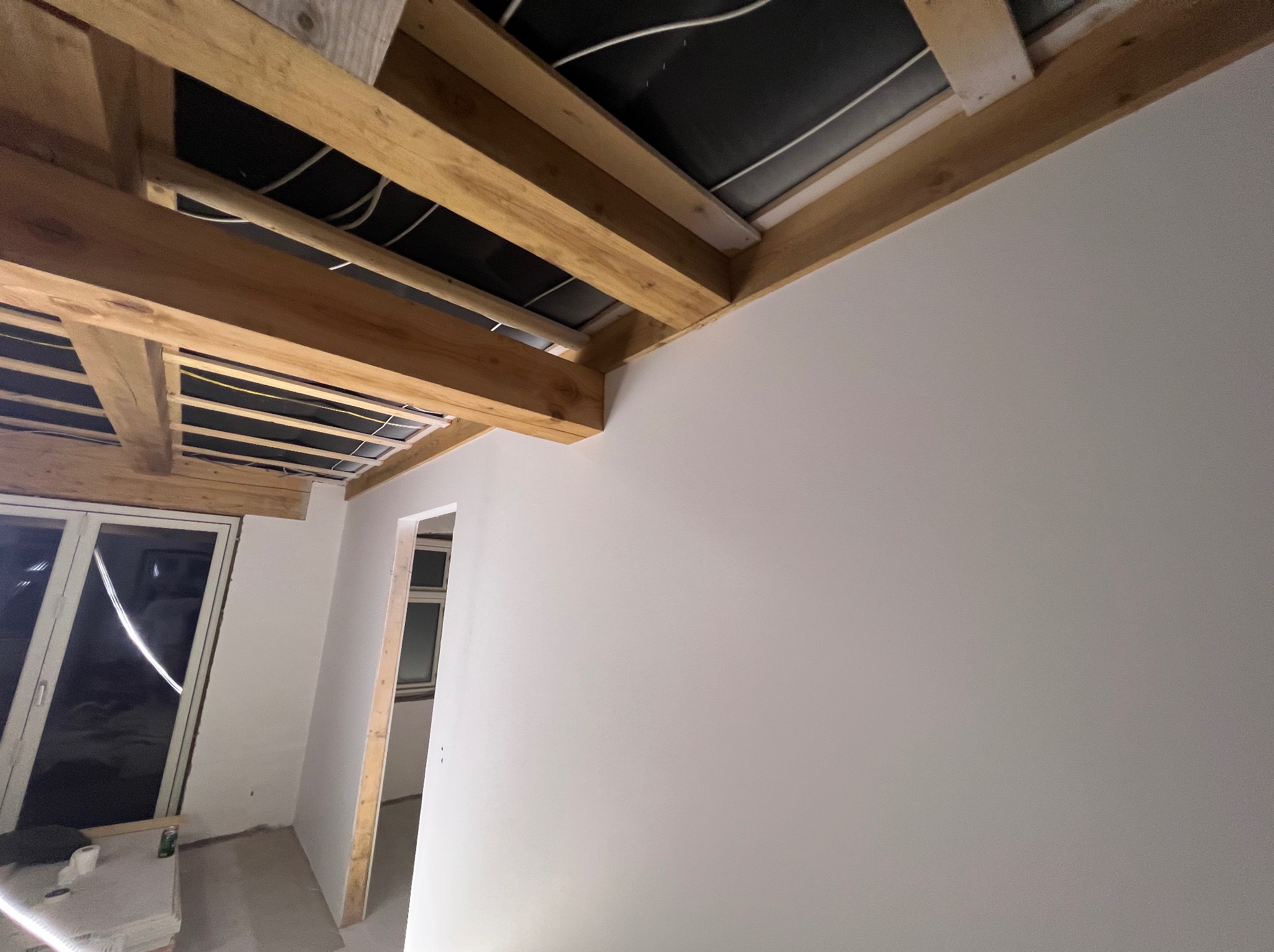 Wnętrze w trakcie prac wykończeniowych: świeżo pomalowana ściana w jasnym odcieniu szarości, widoczne drewniane elementy konstrukcyjne sufitu oraz przygotowane otwory drzwiowe i okienne.