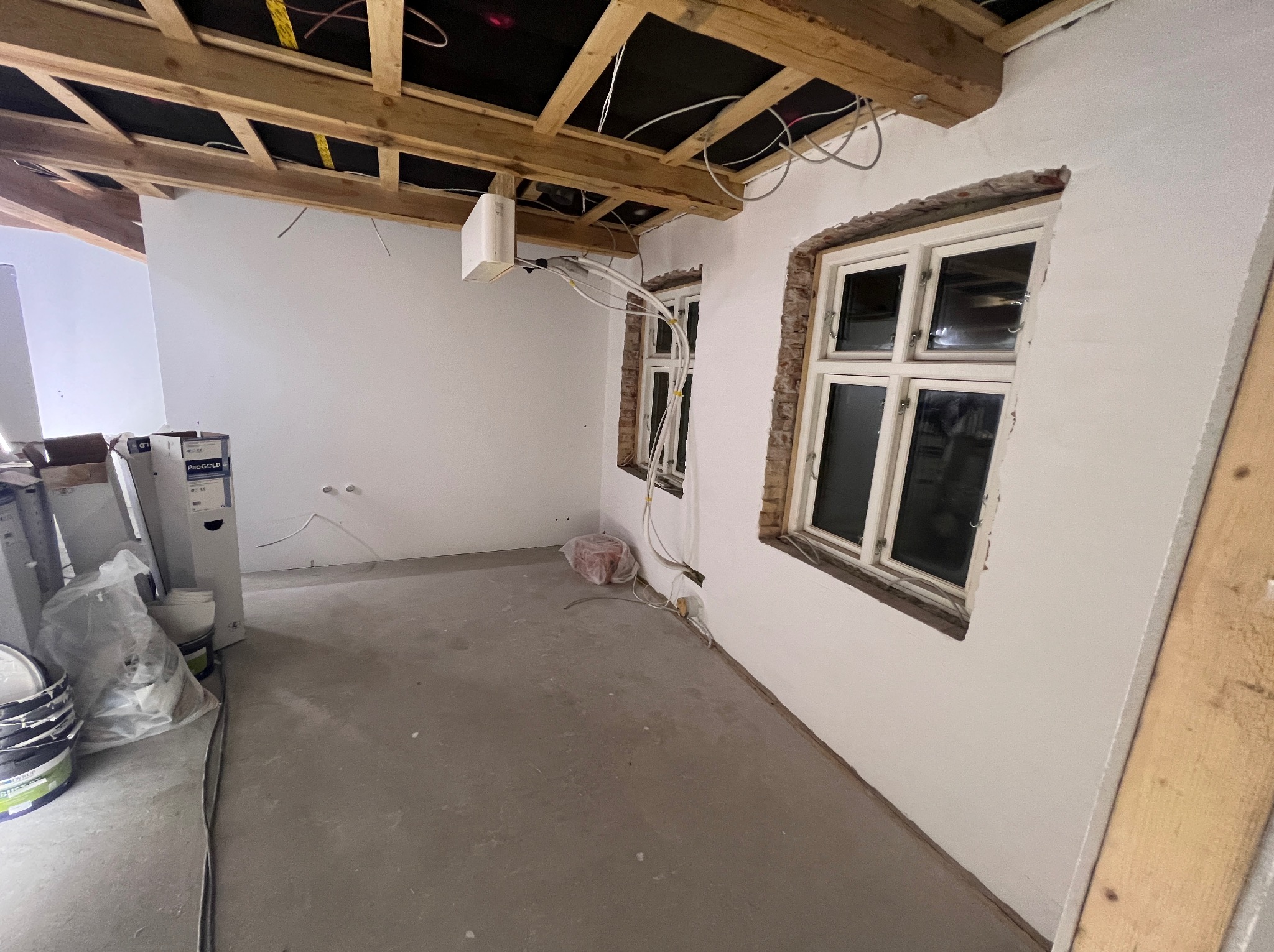 Wnętrze w trakcie remontu z widocznymi oknami po szpachlowaniu, odsłoniętą konstrukcją drewnianą sufitu i przewodami elektrycznymi. Ściany przygotowane pod malowanie.