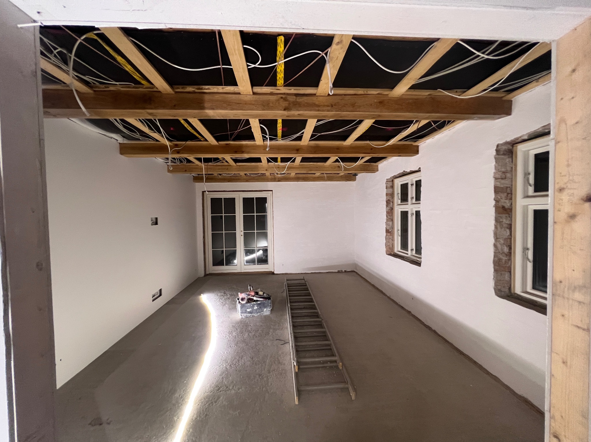 Wnętrze w trakcie remontu w Brzegu: ściany przygotowane do malowania, widoczne drewniane elementy konstrukcyjne sufitu z instalacją elektryczną, drabina i narzędzia na podłodze.