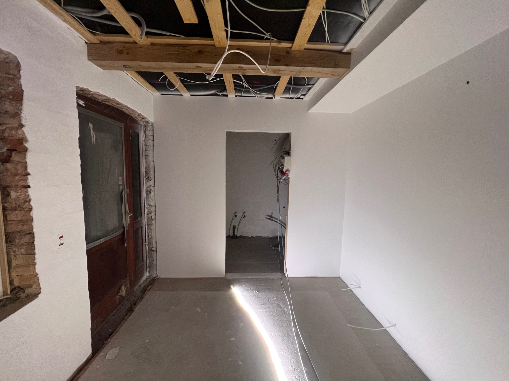 Wnętrze w trakcie remontu: ściany pokryte świeżą, białą warstwą, surowy drewniany strop z widocznymi instalacjami, częściowo odsłonięta cegła przy ościeżnicy drzwi, podłoga przygotowana...