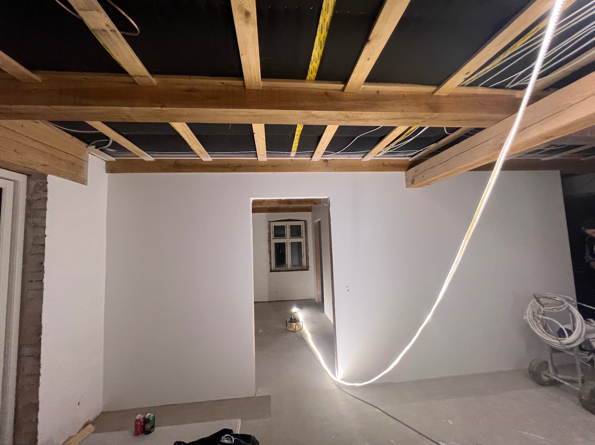 Wnętrze w trakcie remontu: świeżo pomalowane białe ściany, odsłonięty drewniany strop z widoczną instalacją elektryczną, przewód LED na ścianie, sprzęt malarski w rogu, puszki po napojach na podłodze.