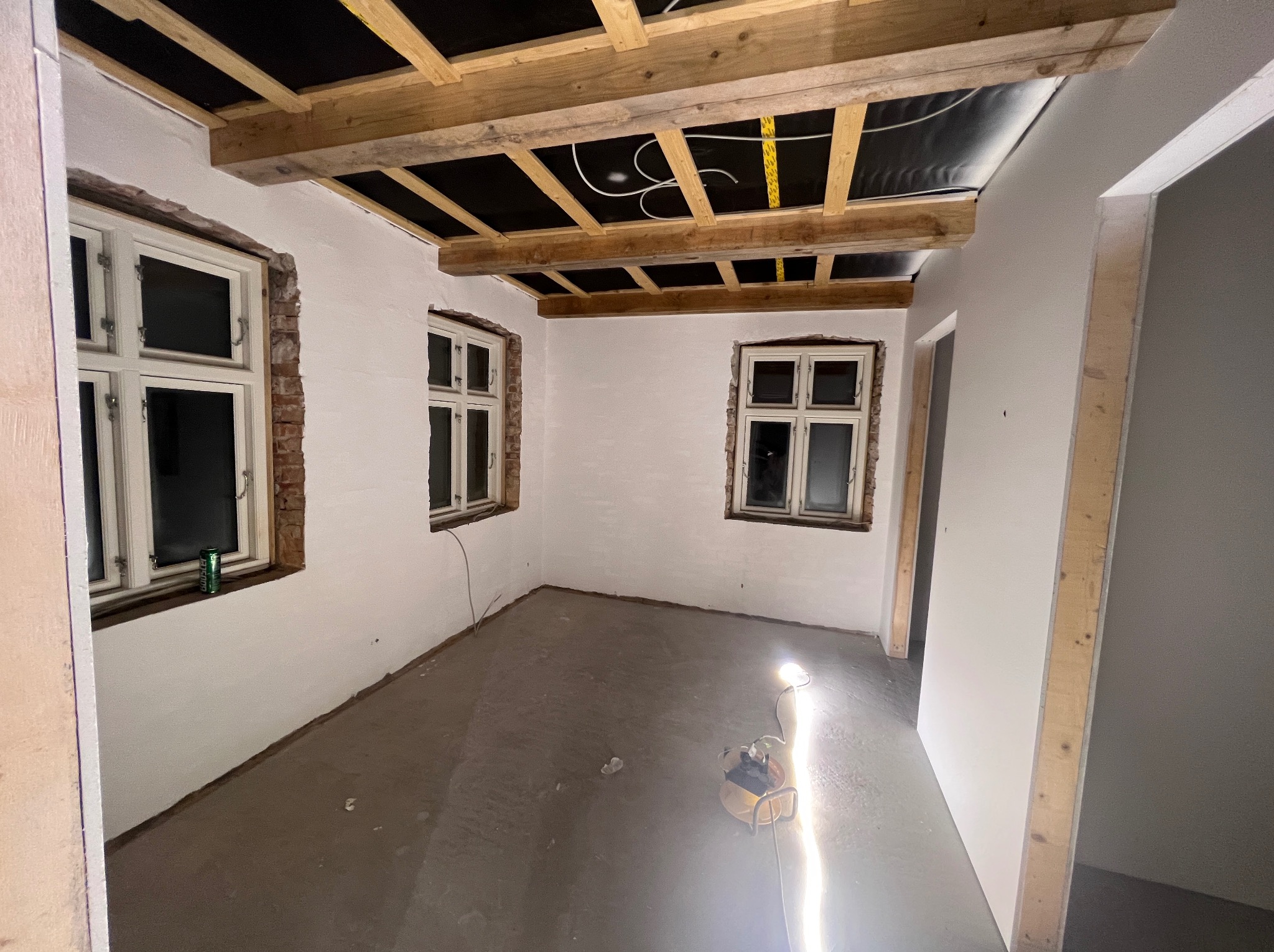 Pokój w trakcie remontu: świeżo położona gładź na ścianach, surowy drewniany strop z izolacją, okna z odsłoniętą cegłą wokół, podłoga przygotowana pod wylewkę, lampa robocza oświetlająca...