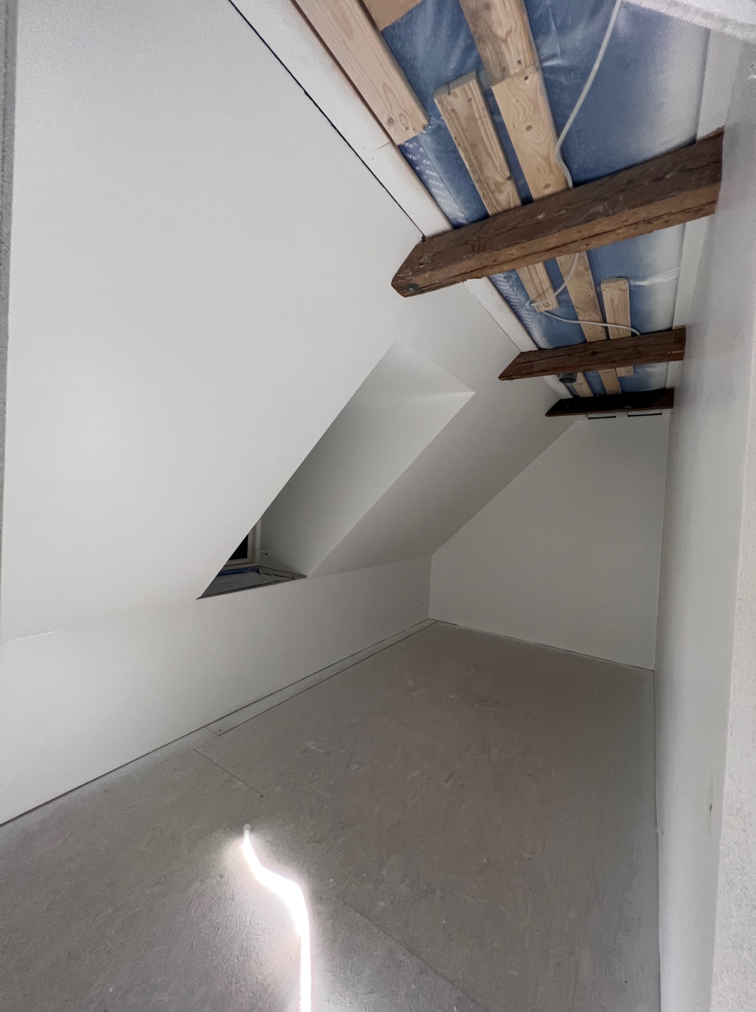 Poddasze po remoncie z gładkimi, białymi ścianami i sufitem, widoczne drewniane elementy konstrukcyjne dachu. Na podłodze widoczny ślad po szlifowaniu.