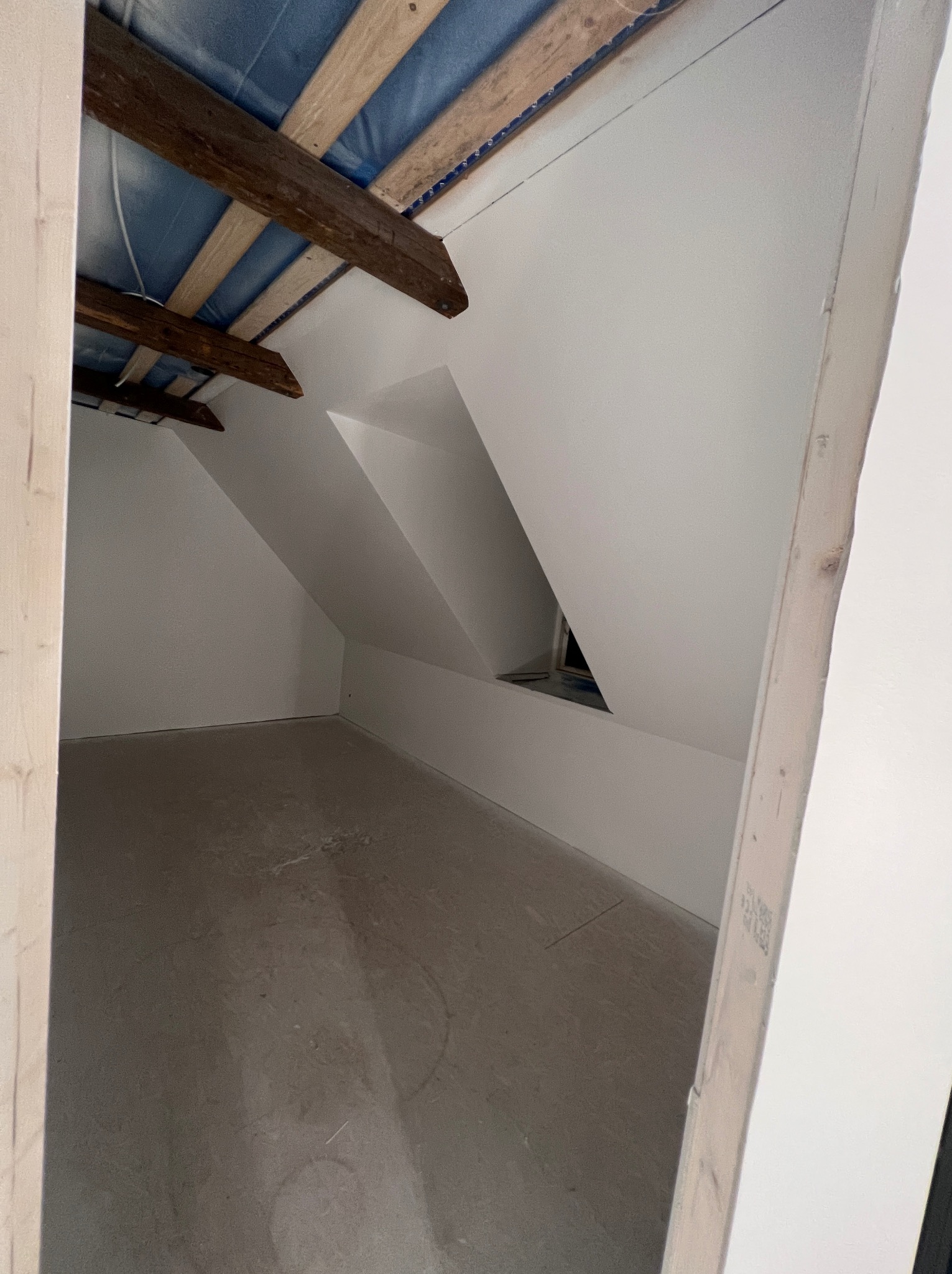 Poddasze po wyrównaniu ścian, widoczne drewniane belki stropowe i okno dachowe w skosie, surowy beton na podłodze.