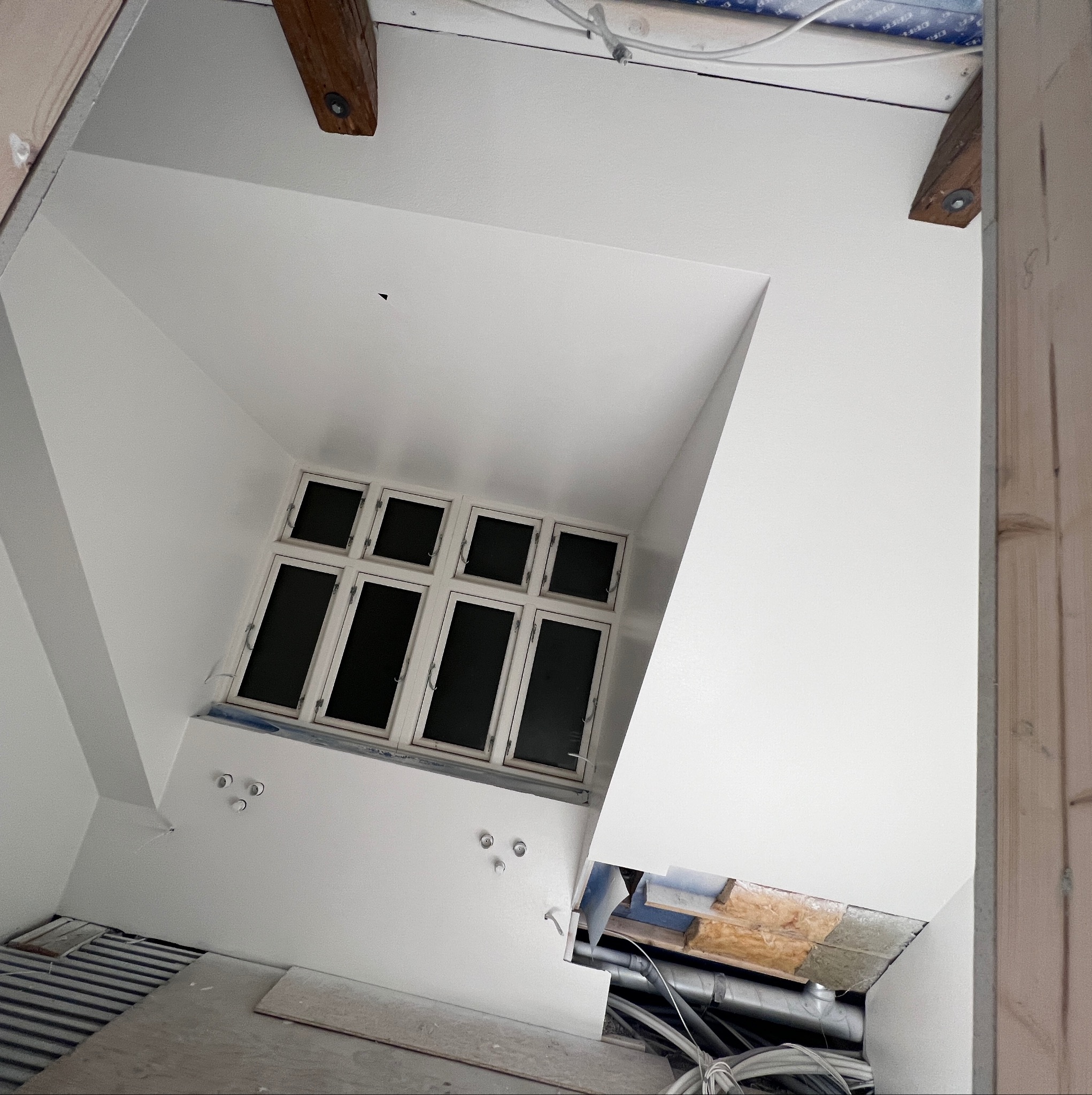 Wnętrze pokoju z oknem po remoncie, świeżo pomalowane ściany i sufit na biało, widoczne drewniane belki stropowe, instalacja elektryczna i fragment podłogi w trakcie prac wykończeniowych.