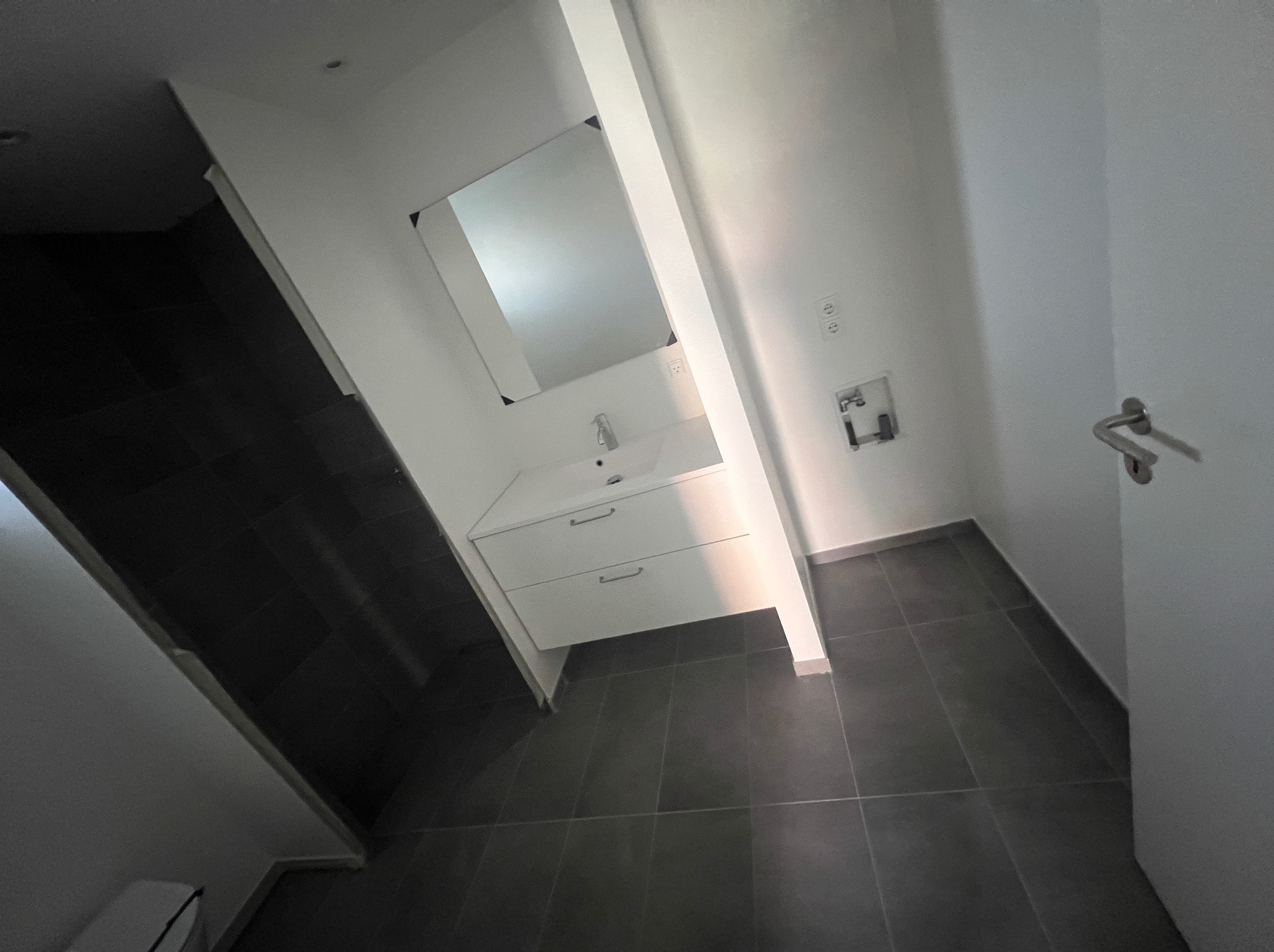 Nowoczesna łazienka z białymi ścianami, szarą podłogą, umywalką z szafką i lustrem, widziana pod kątem, z widocznymi gniazdkami elektrycznymi i drzwiami.