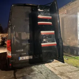 Ciemny bus marki Iveco z otwartymi drzwiami bagażnika, załadowany zapakowanymi w folię stretch przedmiotami zabezpieczonymi taśmą biało-czerwoną, widok z tyłu na numer rejestracyjny DW 6JG07, nocne...