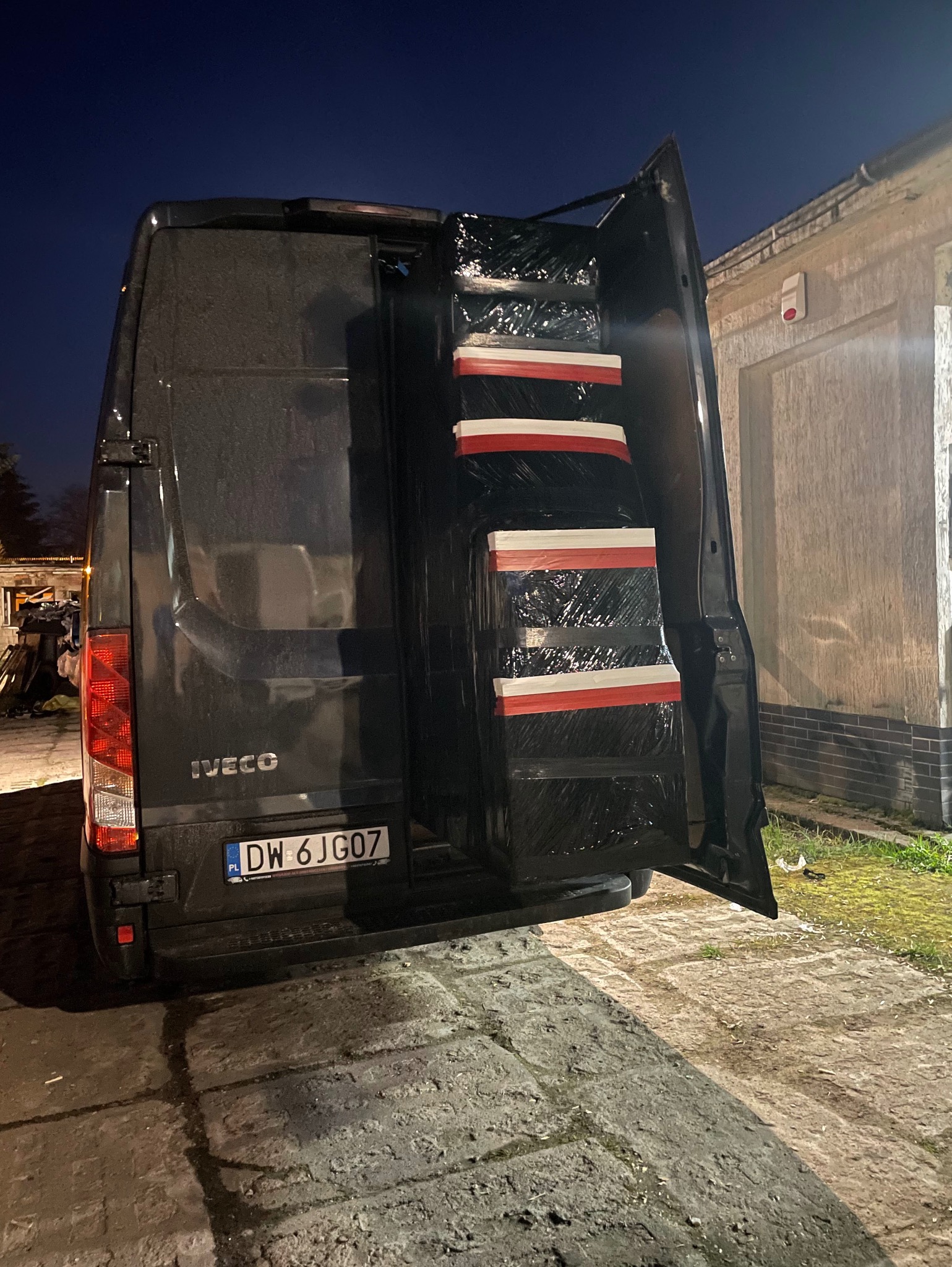Ciemny bus marki Iveco z otwartymi drzwiami bagażnika, załadowany zapakowanymi w folię stretch przedmiotami zabezpieczonymi taśmą biało-czerwoną, widok z tyłu na numer rejestracyjny DW 6JG07, nocne...