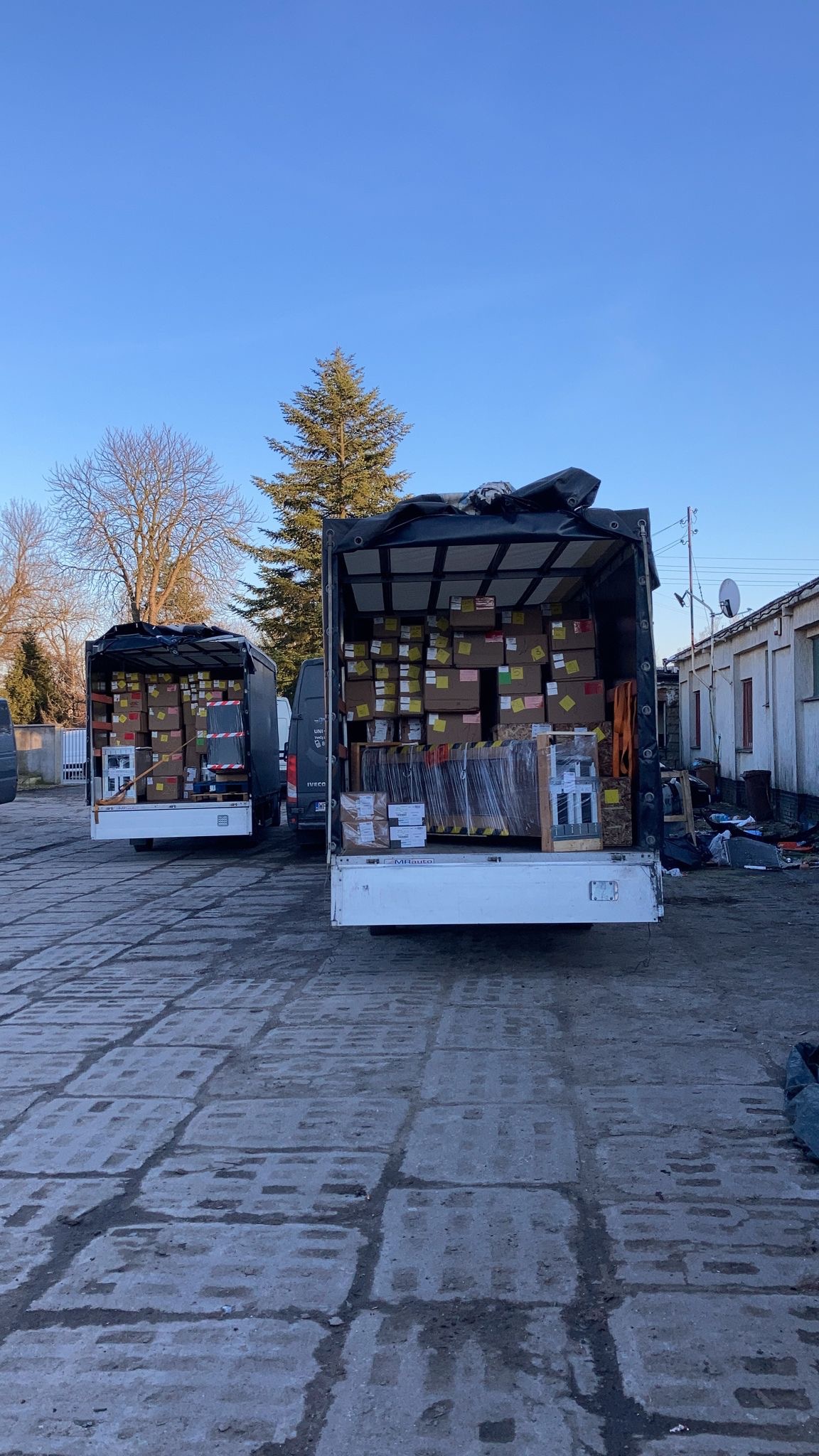 Dwa busy z otwartymi przestrzeniami ładunkowymi, wypełnione kartonami i zabezpieczonymi folią elementami, stoją na betonowym placu w słoneczny dzień. W tle budynek i drzewa.