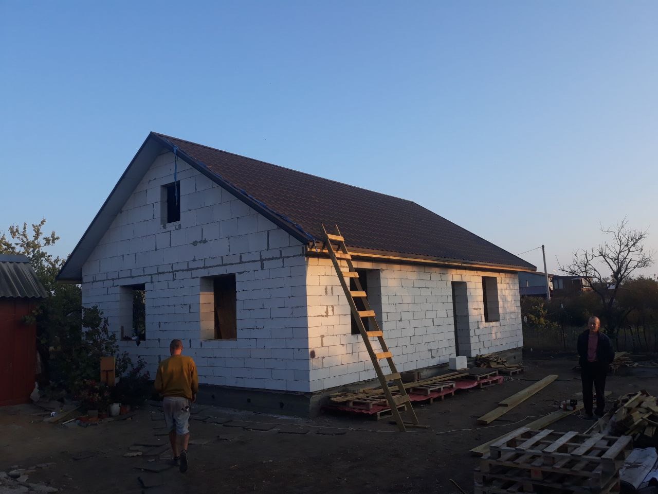 Dom w trakcie budowy z częściowo ukończonym dachem z brązowej dachówki bitumicznej, drabina oparta o ścianę, widoczni dwaj mężczyźni na zewnątrz.
