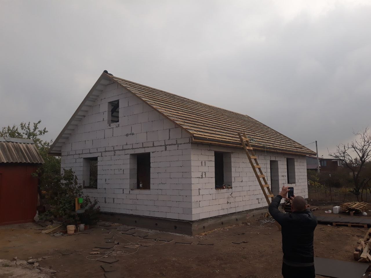 Budowa domu z białych bloczków: widoczne drewniane elementy więźby dachowej, drabina oparta o ścianę, osoba robiąca zdjęcie telefonem.