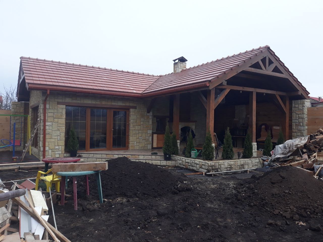 Kamienny dom z drewnianą konstrukcją zadaszenia tarasu, wykończony czerwoną dachówką, widok z zewnątrz na etapie prac wykończeniowych ogrodu.