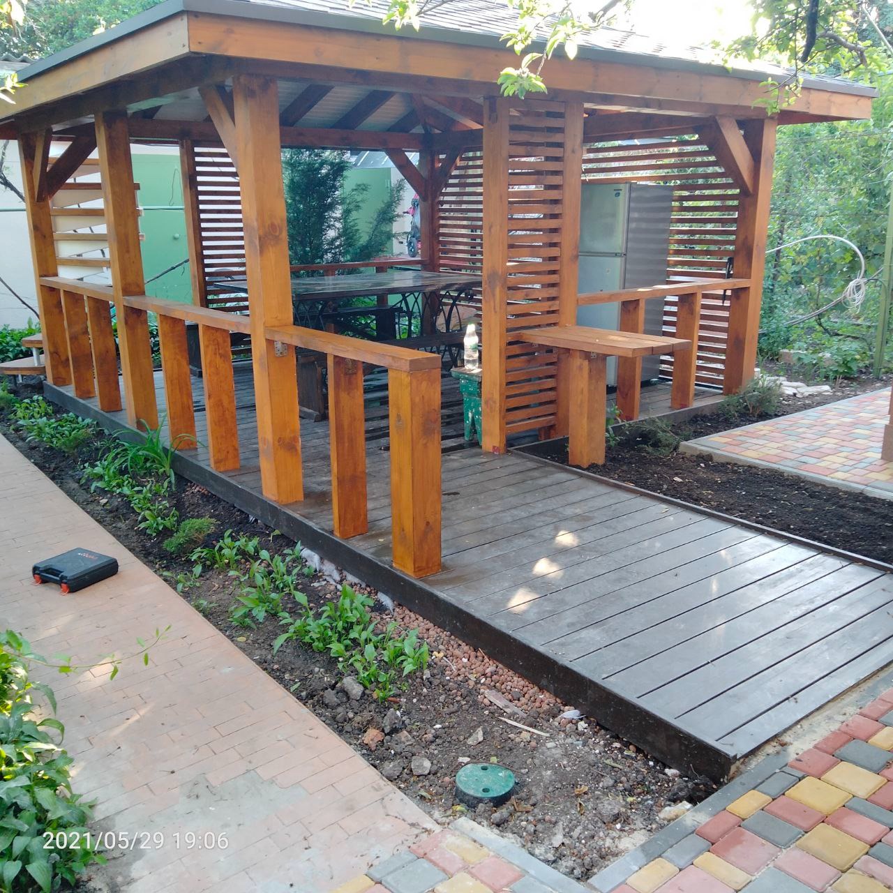 Drewniana altana z zadaszeniem, stolikiem i ławkami, z widoczną więźbą dachową, otoczona zielenią i kostką brukową.