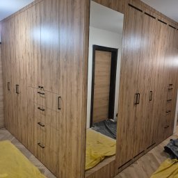 Adam Budzyński - Duża, narożna szafa wnękowa z lustrem i drewnopodobnym wykończeniem, czarne uchwyty, widoczne zabezpieczenia podłogi, nowoczesny design.