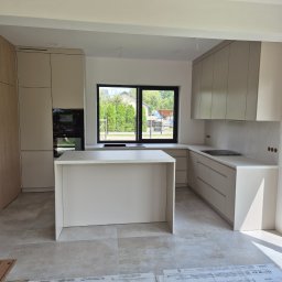 Adam Budzyński - Jasna kuchnia z wyspą i oknem na ogród. Minimalistyczny design, beżowe fronty szafek, płyta indukcyjna. Na podłodze kartony z płytkami Ecoceramic.