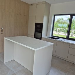 Adam Budzyński - Jasna kuchnia z wyspą i zabudową pod sufit. Okno z widokiem na zieleń. Minimalistyczny design, gładkie fronty, brak uchwytów. Nowoczesne oświetlenie.