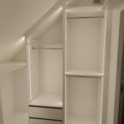 Adam Budzyński - Biała szafa wnękowa na poddaszu z oświetleniem LED, drążkami i szufladami. Widoczna kratka wentylacyjna. Podłoga drewniana. Minimalistyczny design.