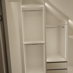Adam Budzyński - Biała szafa wnękowa na poddaszu ze srebrnymi drążkami i oświetleniem LED, dwie szuflady na dole, drewniana podłoga, nowoczesny design.