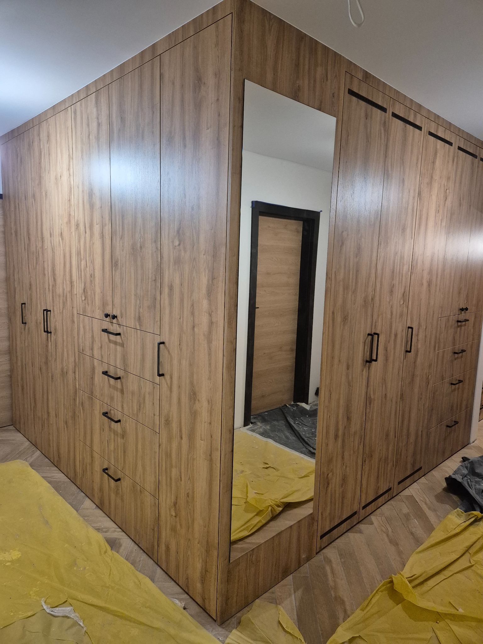 Duża, narożna szafa wnękowa z lustrem i drewnopodobnym wykończeniem, czarne uchwyty, widoczne zabezpieczenia podłogi, nowoczesny design.