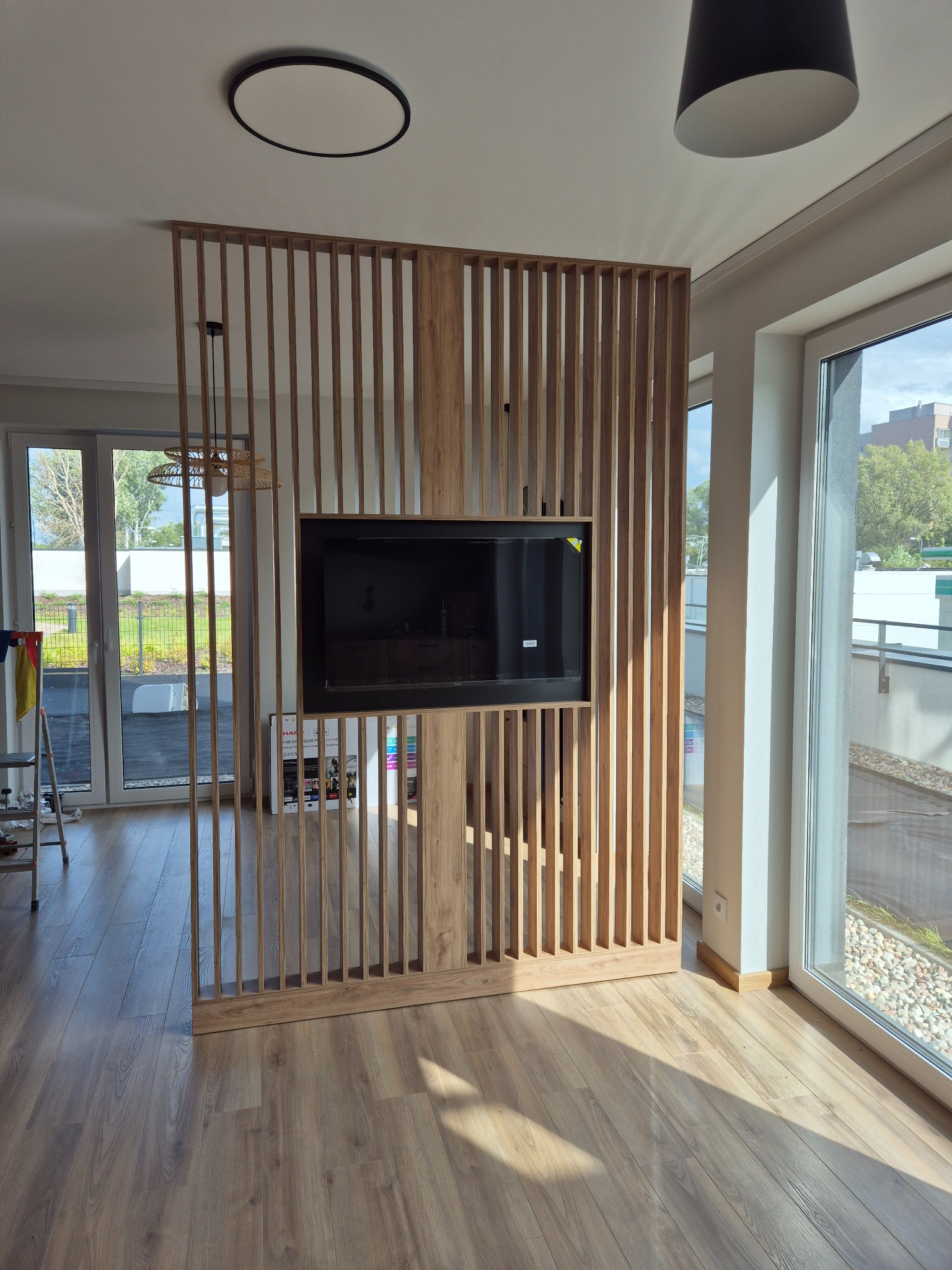 Drewniana ścianka działowa z telewizorem w nowoczesnym salonie. Pionowe lamele tworzą ażurową konstrukcję, w tle widoczne okno i balkon.