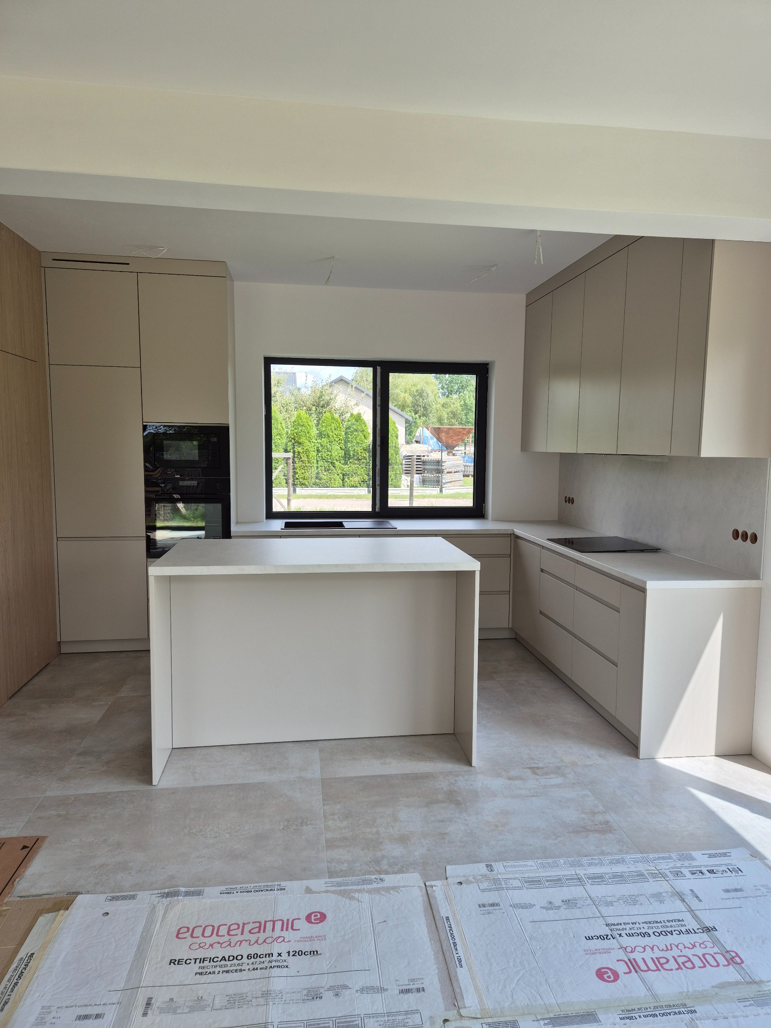 Jasna kuchnia z wyspą i oknem na ogród. Minimalistyczny design, beżowe fronty szafek, płyta indukcyjna. Na podłodze kartony z płytkami Ecoceramic.