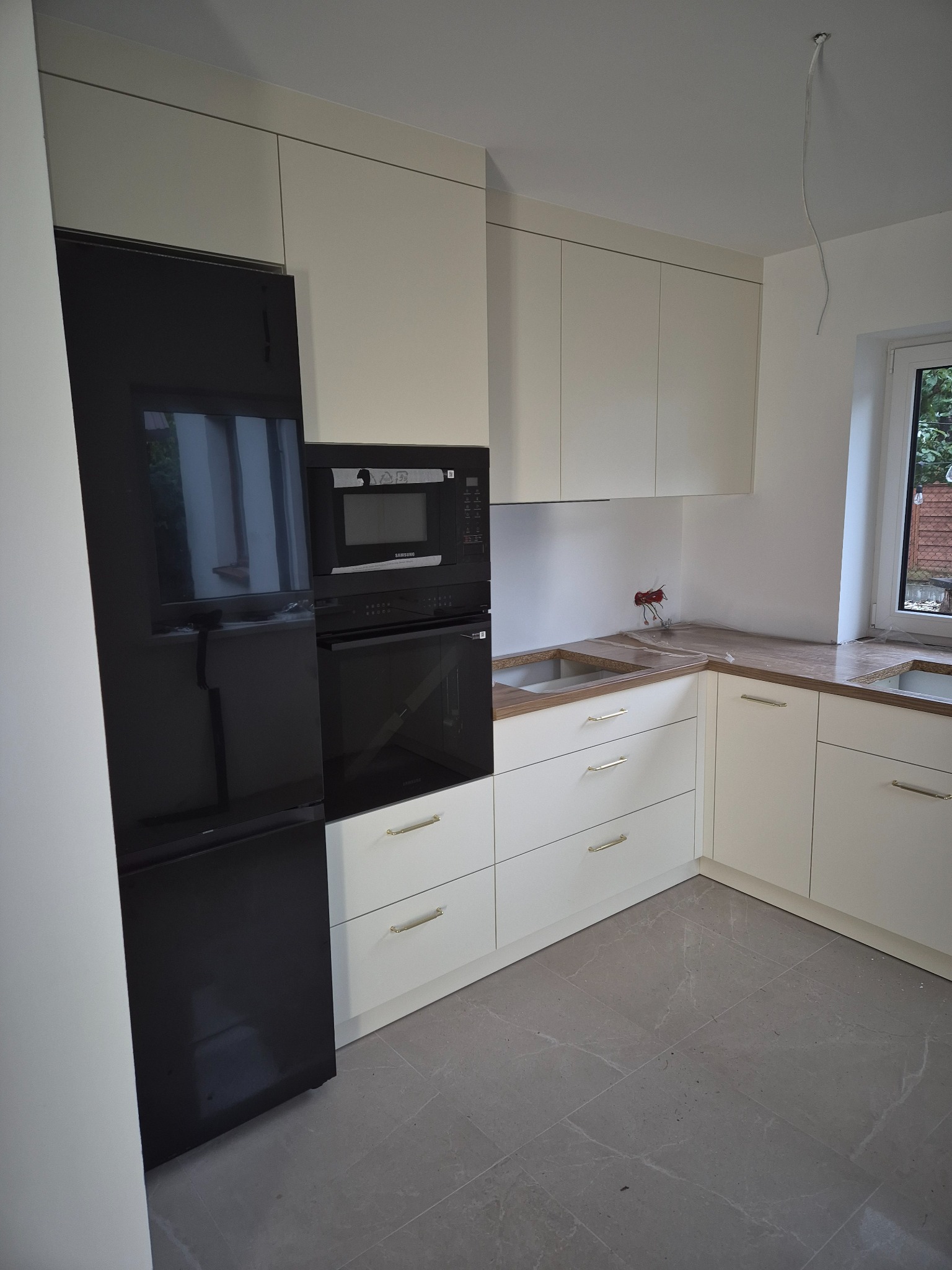 Jasna kuchnia na wymiar z czarnym sprzętem AGD: lodówka side-by-side, piekarnik i mikrofalówka w zabudowie. Blat drewnopodobny, szare płytki podłogowe.