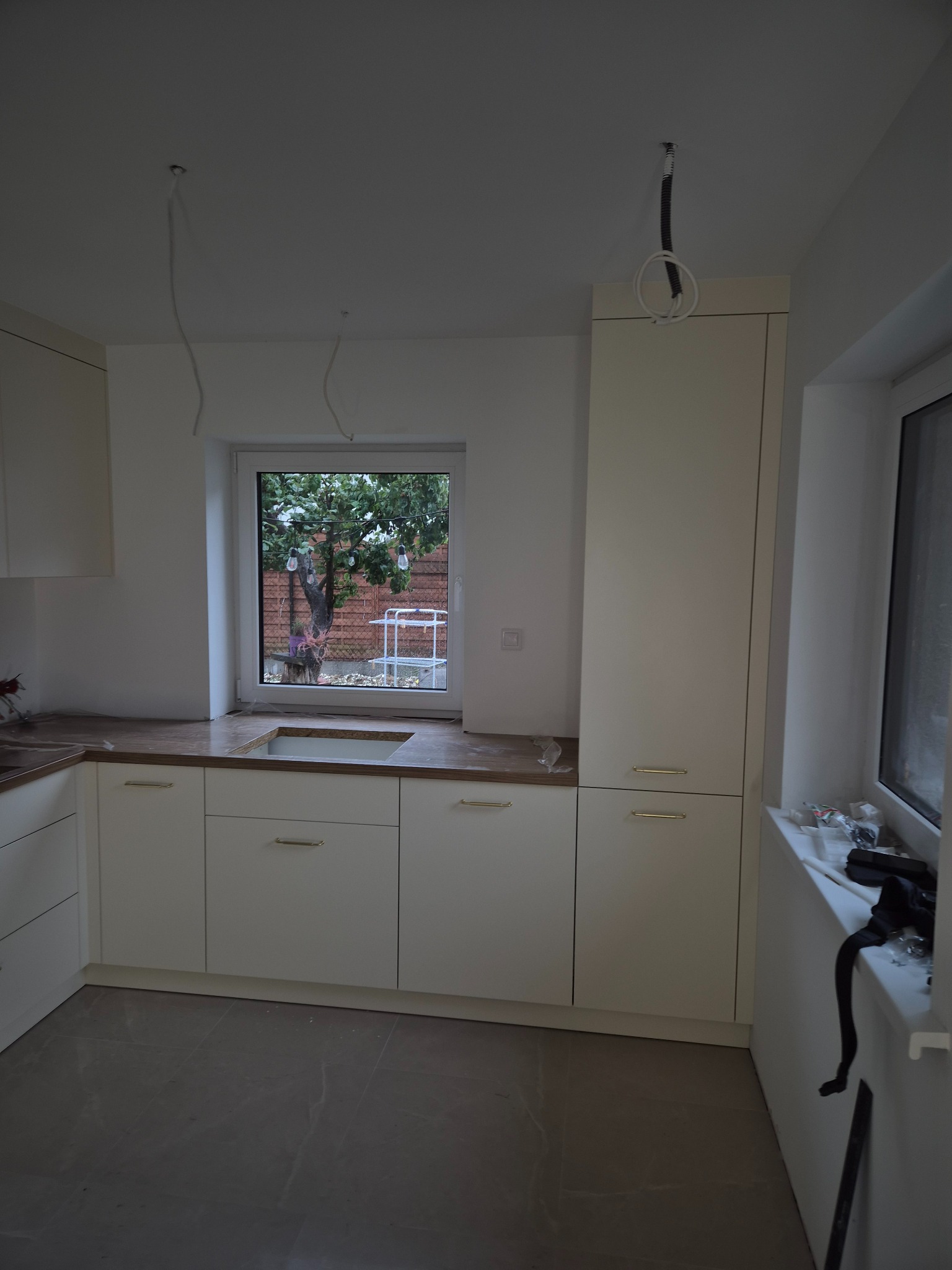 Jasna kuchnia z blatem imitującym drewno, widok na ogród przez okno. W tle widać zwisające kable, co sugeruje etap wykończeniowy.