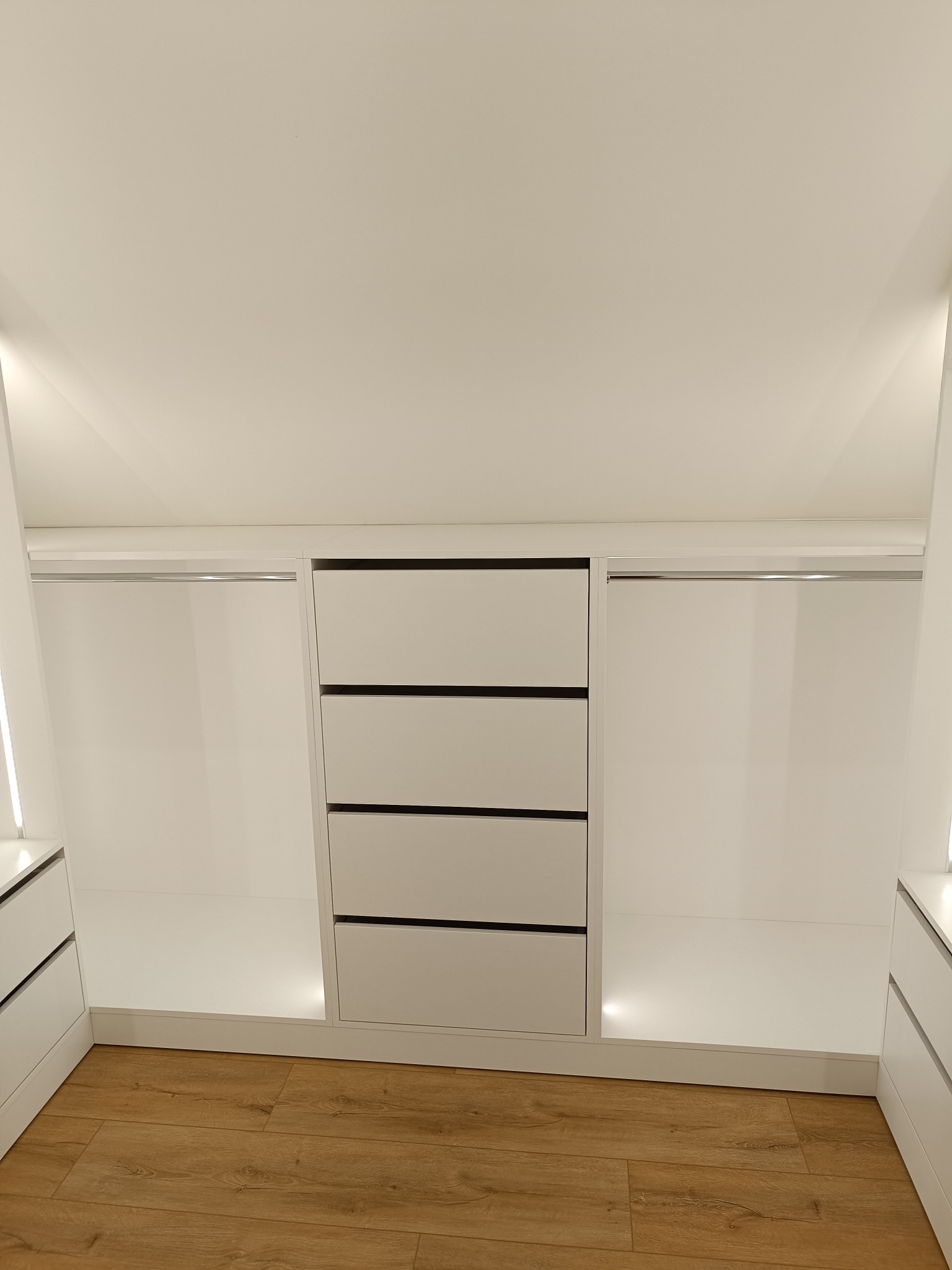 Wnętrze szafy wnękowej z czterema szufladami na środku i drążkami po bokach, oświetlonej od dołu. Jasne drewniane panele podłogowe.