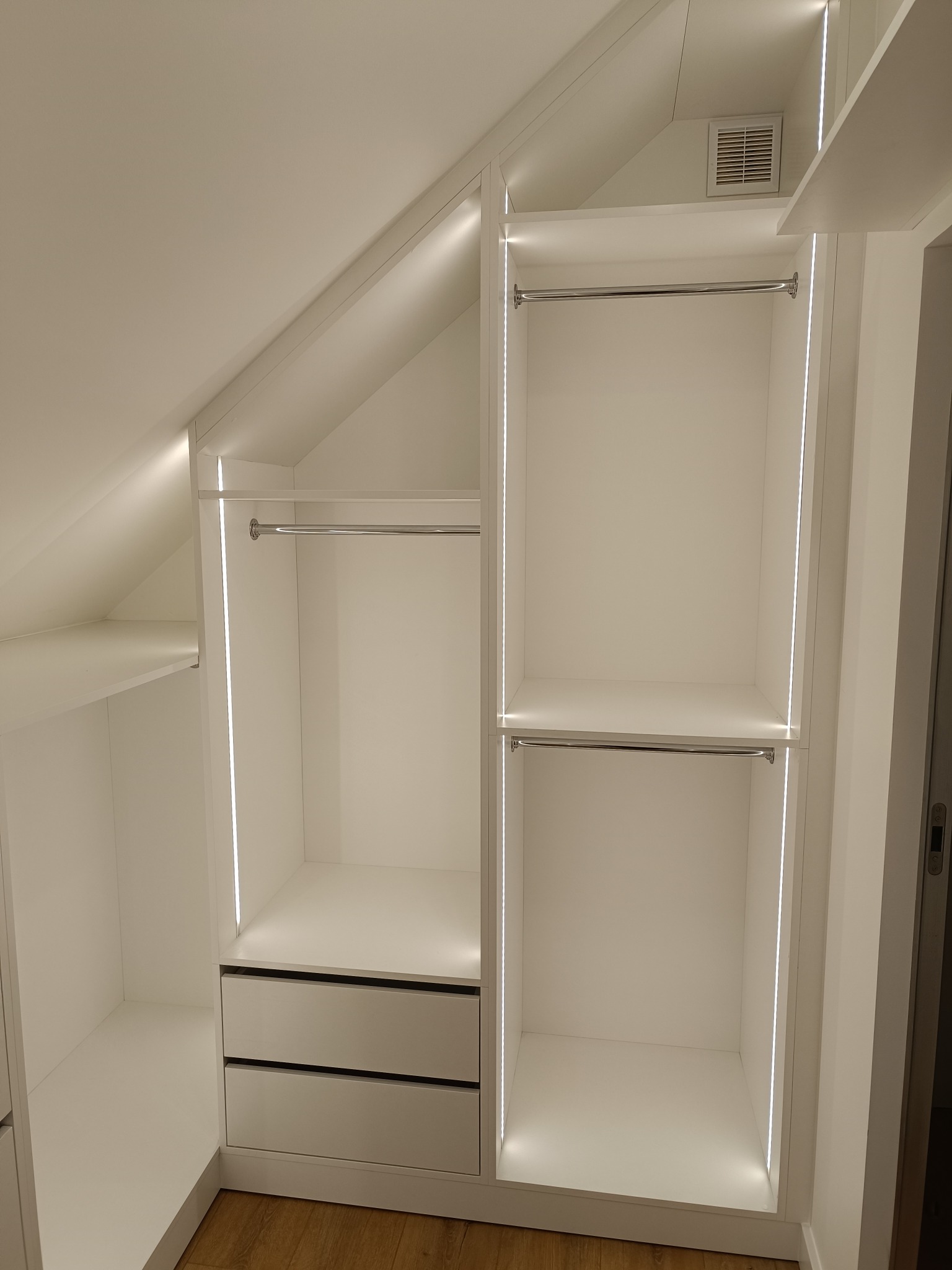 Biała szafa wnękowa na poddaszu z oświetleniem LED, drążkami i szufladami. Widoczna kratka wentylacyjna. Podłoga drewniana. Minimalistyczny design.
