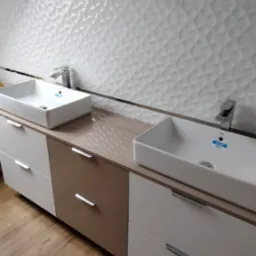 Dwie prostokątne umywalki nablatowe z chromowanymi bateriami, ustawione na szafce z szufladami w kolorach beżu i bieli, tło stanowi biała ściana z trójwymiarową fakturą.