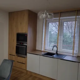 Nowoczesna kuchnia z drewnianymi akcentami: zabudowa kuchenna z piekarnikiem i mikrofalówką, białe szafki podblatowe, czarny zlew z baterią pod oknem z drewnianymi żaluzjami, parkiet na podłodze...