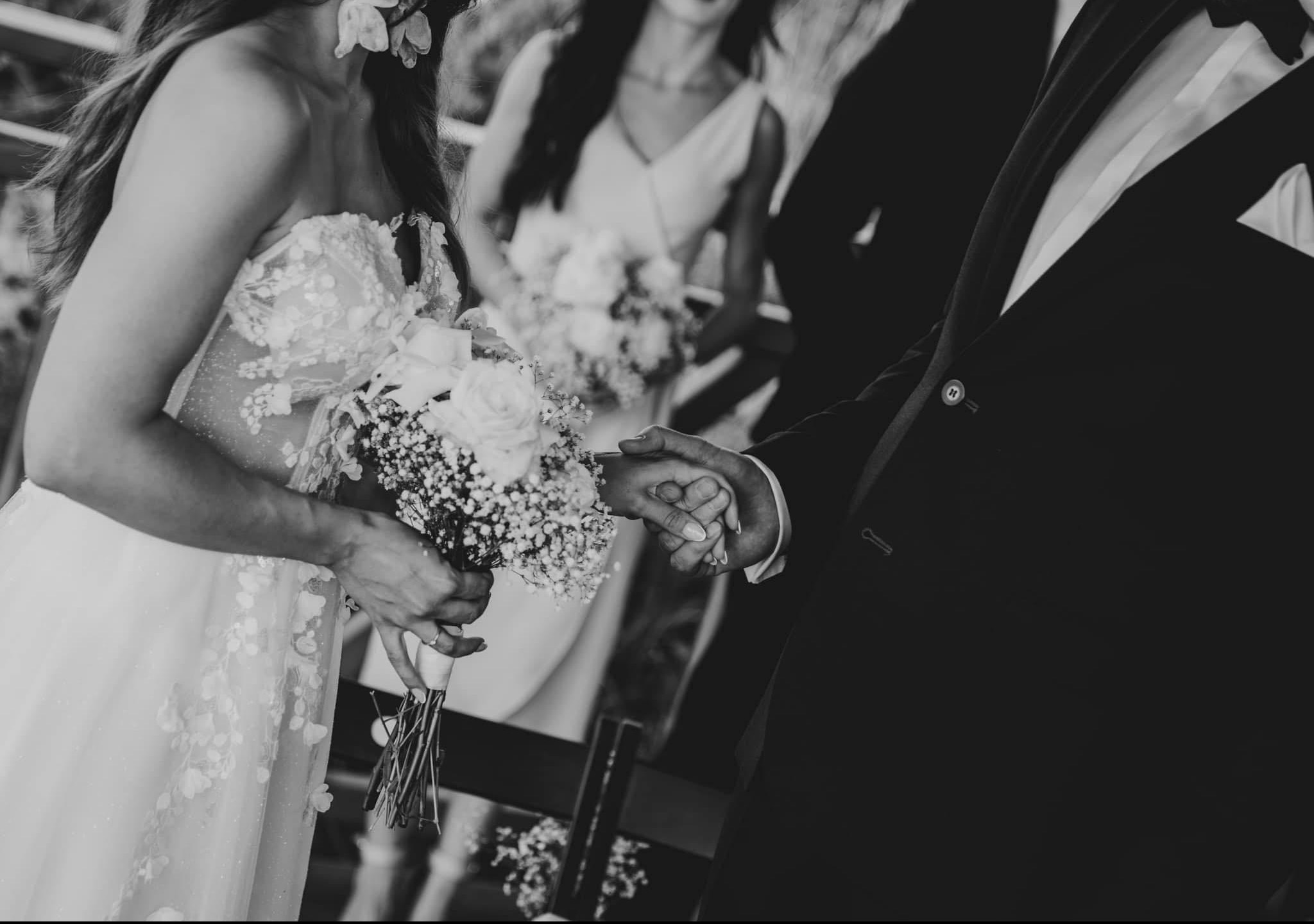 Czarno-białe zdjęcie dłoni nowożeńców splecionych w trakcie ceremonii, panna młoda w sukni z kwiatowymi aplikacjami trzyma bukiet z białych róż i gipsówki, w tle druhna z bukietem.