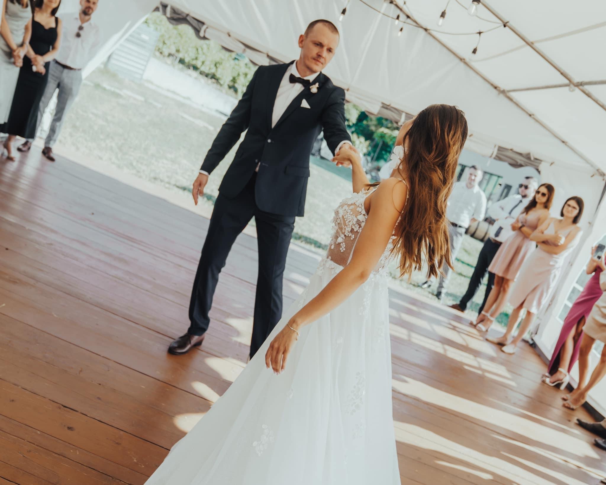 Para młoda tańczy pierwszy taniec w namiocie udekorowanym lampkami, goście obserwują w tle.