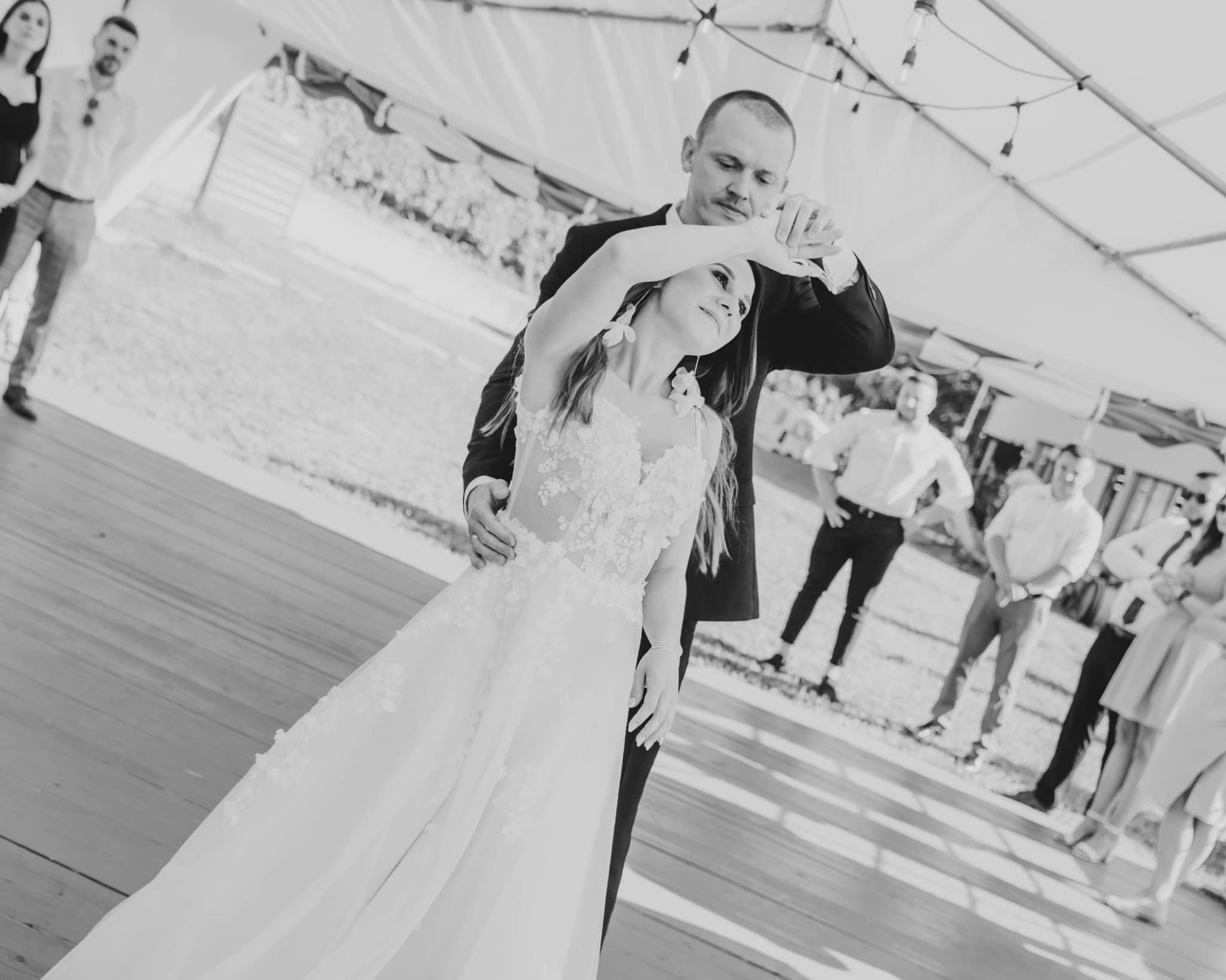 Czarno-białe zdjęcie pary młodej tańczącej pod namiotem, panna młoda w sukni z kwiatowymi aplikacjami i dużymi kolczykami, pan młody w ciemnym garniturze, w tle goście.