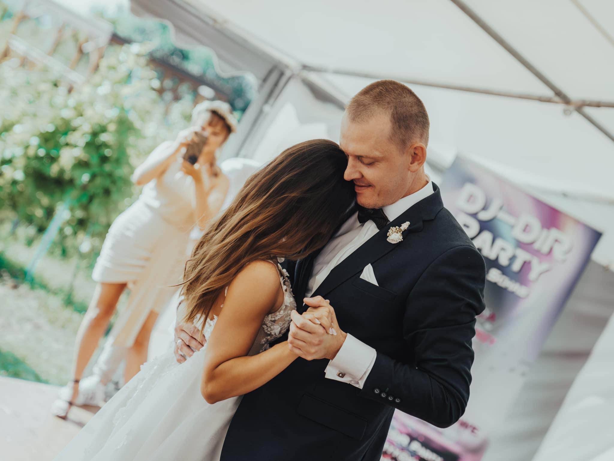 Para młoda w tańcu podczas wesela na zewnątrz, panna młoda w białej sukni z koronką, pan młody w czarnym garniturze z muszką, w tle osoba robiąca zdjęcie telefonem i plakat DJ-a.