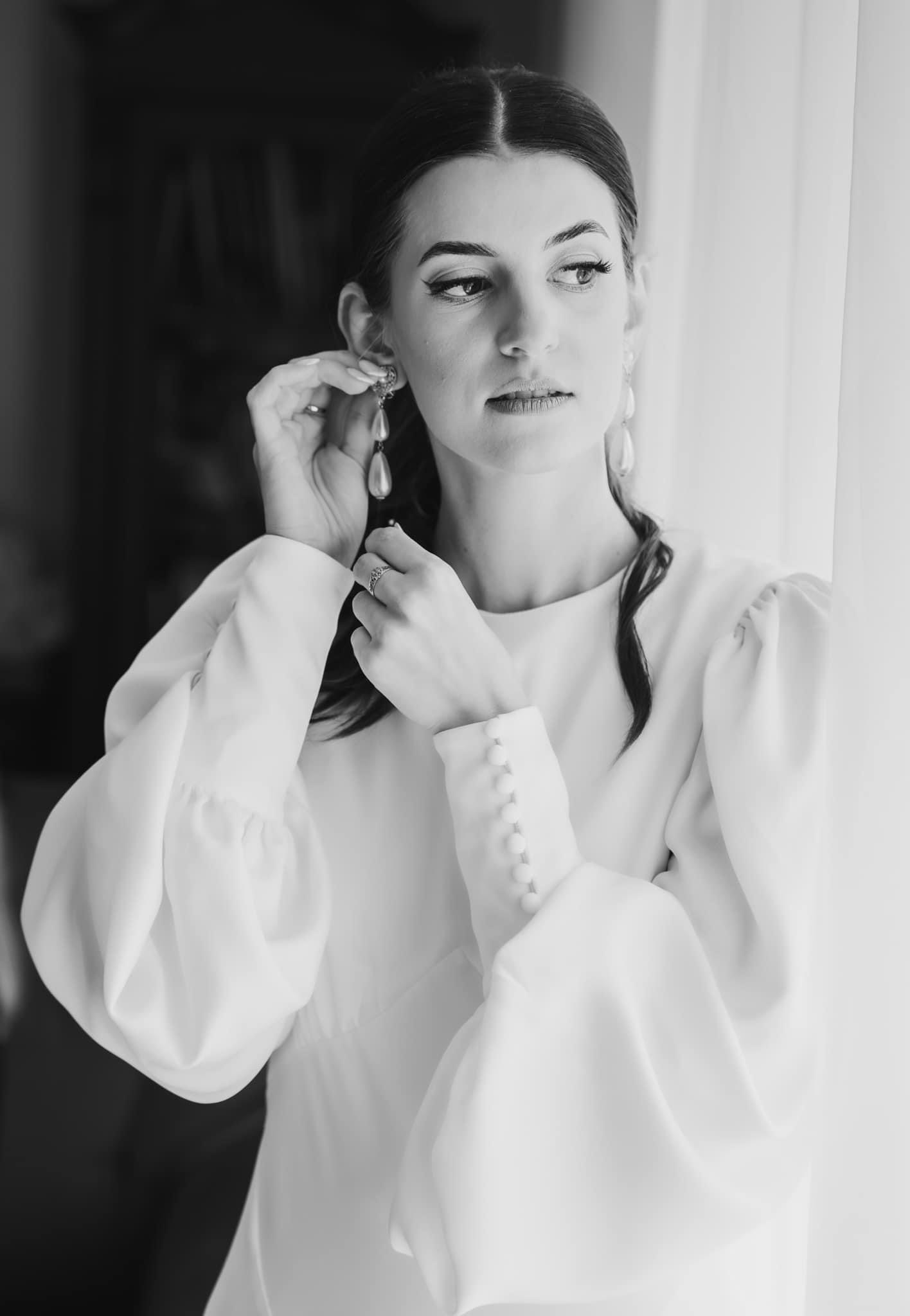 Czarno-białe zdjęcie panny młodej zakładającej kolczyk z perłą, z pierścionkiem zaręczynowym na palcu, w białej sukni z bufiastymi rękawami z rzędem małych guzików, spoglądającej w bok na jasne okno.